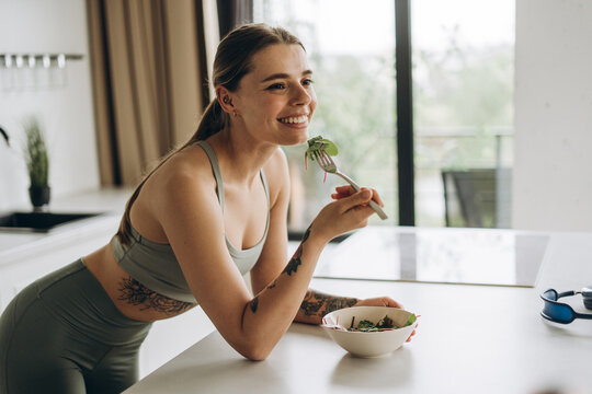 Sportswoman, young woman wearing stylish sportwear eating salad in the kitchen after workout