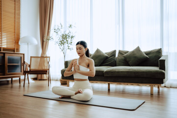 Young Asian woman meditating in lotus position at home, sitting on the floor in fitness clothing. Breathing workout to achieve relaxation, peace and mindfulness