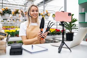 A Caucasian woman, working in a florist shop, presents gardening tools while streaming online from a table with a laptop and smartphone.