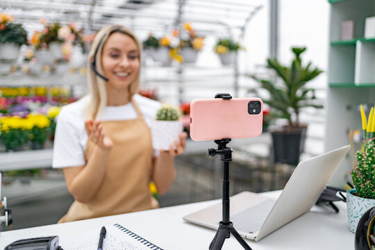 A Caucasian woman in a florist shop hosts a live sale of a cactus, using her smartphone for a broadcast.
