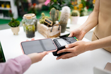 A customer pays with a phone using contactless payment at a florist shop. Various plants are...