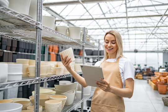A smiling florist examines a flowerpot in a shop, using a tablet while surrounded by an array of plant containers on shelves.