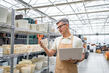 A Caucasian man inspects a flowerpot in a plant shop, holding a laptop for inventory. The shop is...