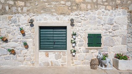 A charming Mediterranean stone house facade with green wooden shutters, rustic outdoor lighting, decorative flower pots, and a tree stump stool.