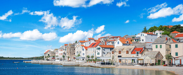 Fototapeta premium A stunning coastal town of Primosten in Croatia features traditional stone houses with red roofs beside the crystal-clear Adriatic Sea under a vibrant blue sky.