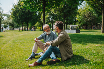 Father and adult son sitting on grass in park, sharing a story from the past.