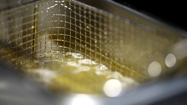 Close-up of bubbling oil in industrial deep fryer, metal basket partially submerged, golden light.