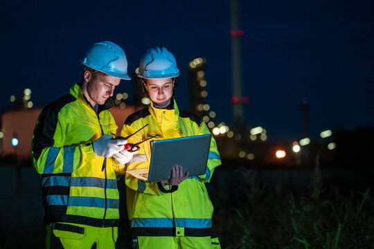 Industrial teamwork of engineer man and woman using a laptop computer during night shift at an oil refinery. Focused professionals working under factory lights.