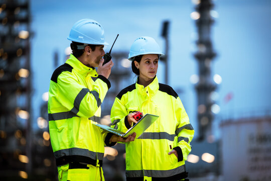 Factory worker engineer man and woman using laptop at night, working together on refinery site with industrial lights in background. Professional teamwork at night shift.