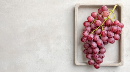 Bunch of red grapes in a square white ceramic tray. the grapes are bunched together and appear to be fresh and ripe. the tray is placed on a light grey textured background.