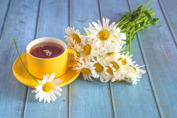 Yellow cup of tea and bouquet of white daisies flowers on blue wooden background; close up