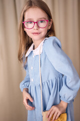 A beautiful young girl in a blue dress with a white collar and glasses coquettishly adjusts a small straw bag with her hand. She has long blond hair and looks at the camera with a slight smile. 