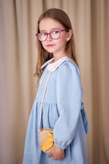 A charming little girl with a cute wicker handbag over her shoulder stands half-turned on a milky fabric background. The child in glasses is dressed in an elegant velvet dress with a white collar