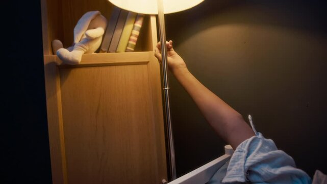 Tilt down shot of curious little Black girl in pyjamas lying in bed under blanket at night in dark room and playing with floor lamp, turning it on and off