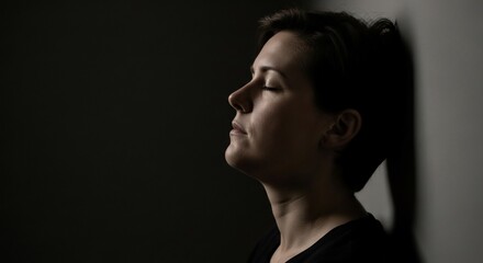 Contemplative woman with closed eyes leans against a wall in shadow
