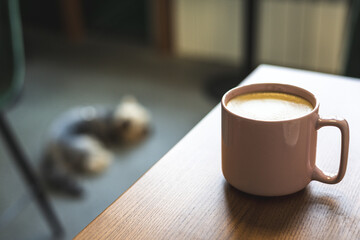 A cozy morning scene with a pink coffee mug and a blurred dog. A cup of coffee on a wooden table, with a dog lying in the background. Inviting scene.