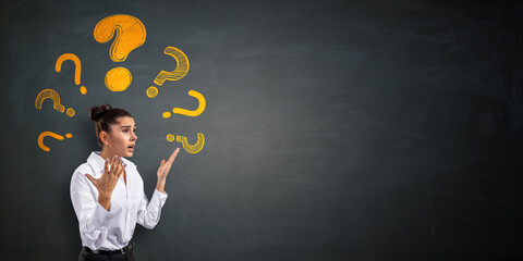Confused businesswoman surrounded by yellow question marks on a dark chalkboard background with...