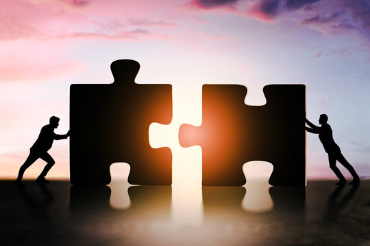 Silhouettes of businesspeople pushing giant puzzle pieces together at sunset to represent teamwork and collaboration in a business concept.