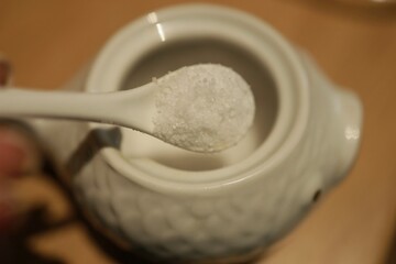 A hand holds a white spoon with coarse salt, poised above a finely designed container on a wooden...