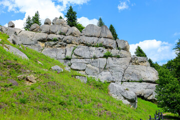 Mighty rock formations, resembling ancient walls, rise above a green slope overgrown with grass and small shrubs. Some of them have unusual protrusions, creating the impression of architectural elemen