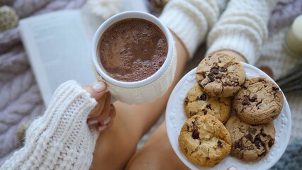 Cozy winter scene with a woman relaxing on a bed, holding a mug of hot chocolate and a plate of cookies, with an open book nearby, creating a warm and inviting atmosphere