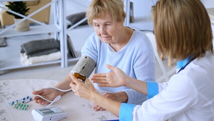 Female doctor holding medicine bottle is explaining prescription to senior woman while measuring blood pressure with automatic blood pressure monitor