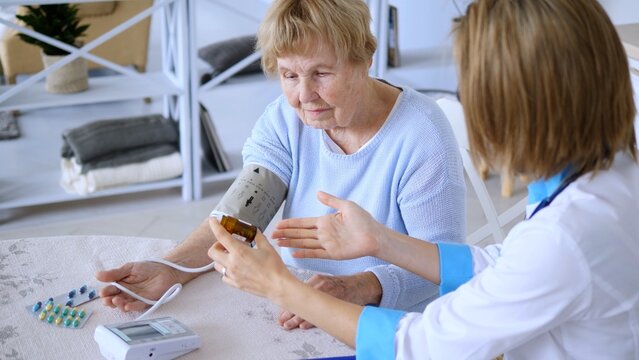 Female doctor holding medicine bottle is explaining prescription to senior woman while measuring blood pressure with automatic blood pressure monitor
