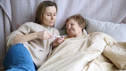 Young woman supporting her sick elderly mother lying in bed, checking her temperature with a...
