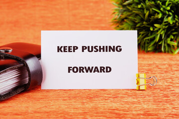 Concept text KEEP PUSHING FORWARD written on a card on a bright orange table next to a black...