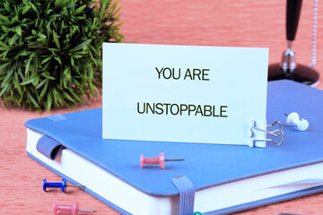 Concept text YOU ARE UNSTOPPABLE written on a card on a blue notebook next to colorful pins and an...