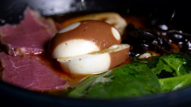 Close-up of ramen bowl with soy-marinated egg, thin beef slices, spinach, and dark mushrooms in broth.