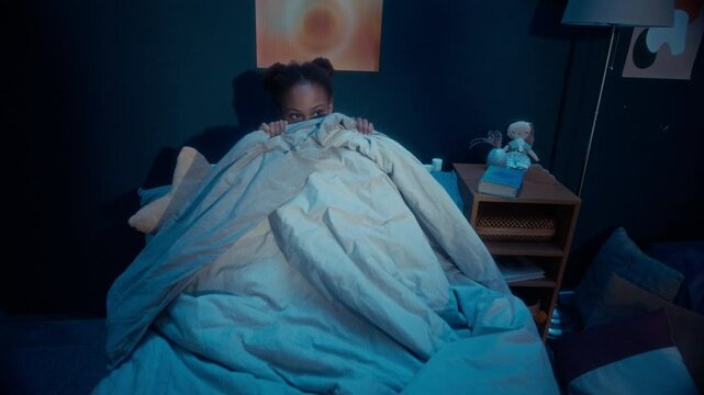 Frightened African American little girl covering herself under blanket sitting on bed in dark kids bedroom with creepy hand silhouettes playing shadow game on wall