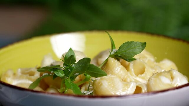 Close-up of cheesy shell pasta with green basil leaves on top, served in a vibrant yellow ceramic bowl.
