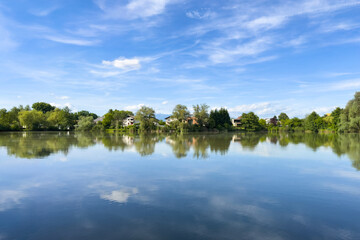 a serene lake reflects a clear blue sky and lush greenery with houses among trees. nature symmetry and contrast. minimalist