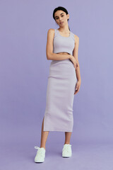 young woman in lilac ribbed dress standing against purple background with casual sneakers,...