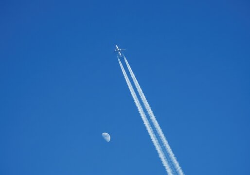 Airplane flying in blue sky with contrails and the moon visible