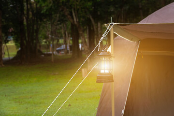 close-up of a black camping lantern hanging under a tarp in a lush green outdoor setting. The LED lantern is suspended by a rope. The background shows trees and grassy ground