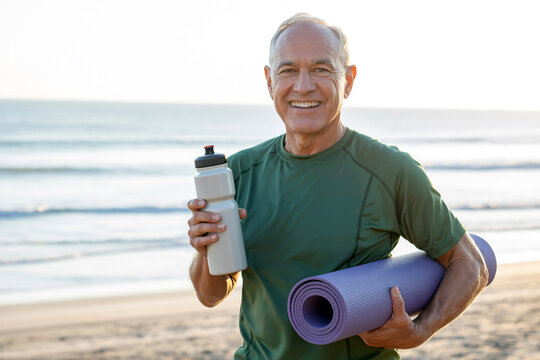 A smiling senior man stands on a beach holding a water bottle and yoga mat