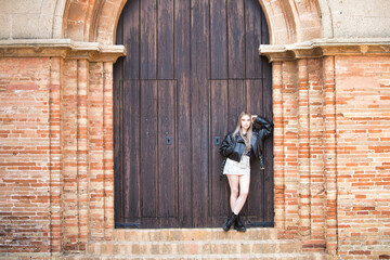 Young and beautiful blonde woman in black leather jacket, boots and short skirt leaning on the...