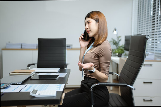 Asian businesswoman talking on mobile phone while working on laptop at office desk. communication, and corporate success.