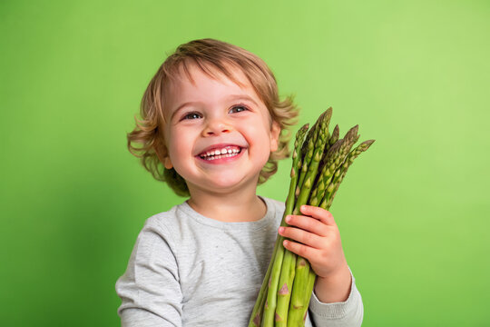 Cute kid holding asparagus vegetable and smiling over green studio background. Healthy relationship with food and child nutrition concept. Healthy baby food with vitamins and kids vegetarianism.