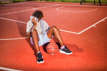 Young black woman is sitting on basketball court. She is tired and wiping sweat of her face.