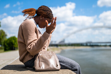 Portrait of displeased young black woman who is sitting by the river in the city.