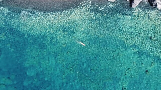 4k video drone top down view of a woman swimming in a breath taking turquoise beach, revealing the rocky cliffs and stunning scenery