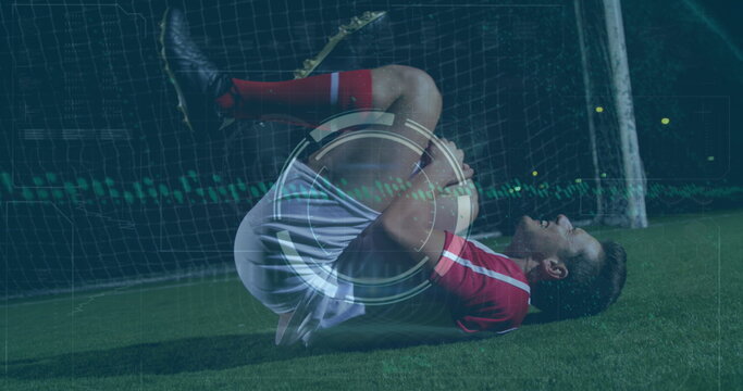 Lying soccer player in red uniform clutching knee on grass pitch by floodlights, with goal net