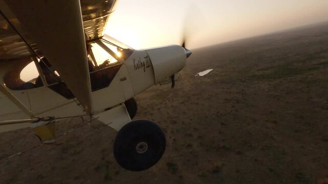 Wide from side of small light plane flying over savannah with shadow casting and aerial of sparse vegetation at golden sunset in Kenya