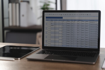 Report file laptop displaying spreadsheet with data and charts wooden desk modern office environment, with tablet and pen nearby, creating