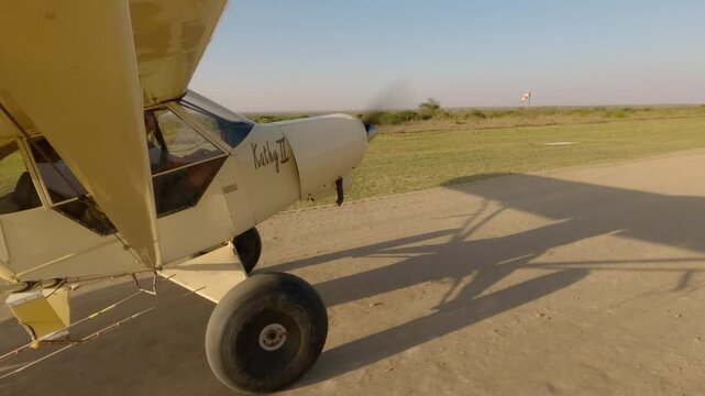 Wide side view of two person small light plane taking to flight off of dusty dirt runway in Savannah on clear sky sunny day in Kenya