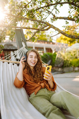 Stylish woman with wireless headphones and phone relaxing lying in hammock in sunny park. Beautiful...