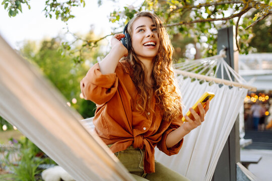 Stylish woman with wireless headphones and phone relaxing lying in hammock in sunny park. Beautiful woman enjoying listening to music or podcast outdoors. Relaxation and technology concept.